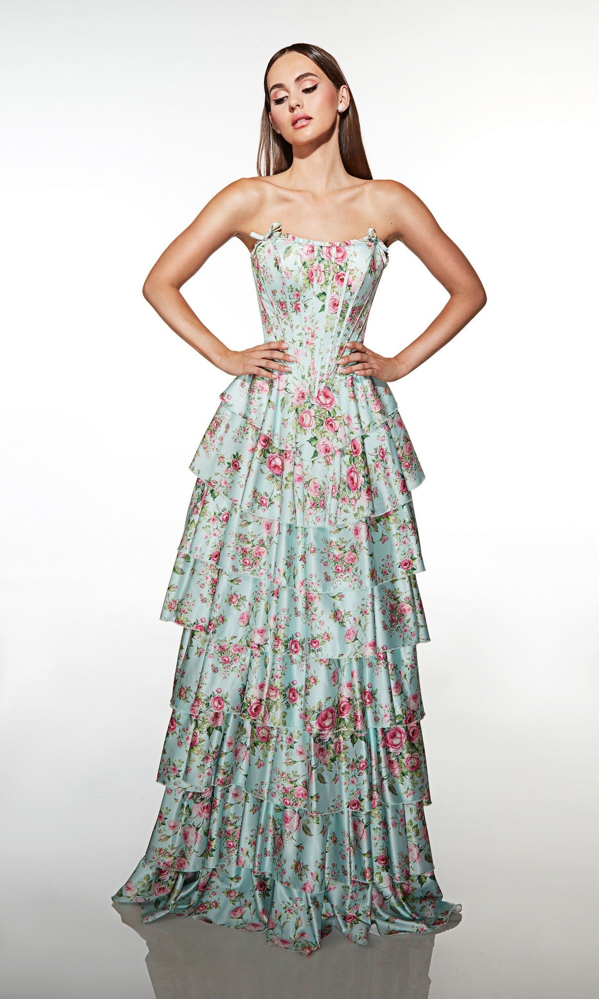 A woman poses with hands on hips, wearing the Alyce 62066 long prom dress—strapless, light blue with pink florals, featuring a corset bodice and tiered ruffle skirt—against a plain white backdrop.